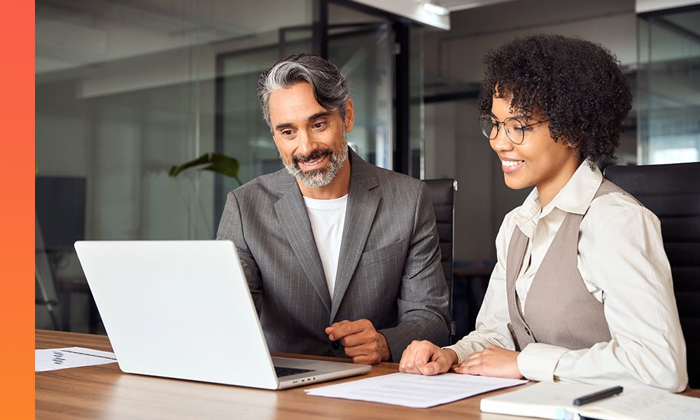 Two professionals sit at a table and smile looking at a laptop.
