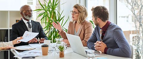  Three people sitting in a conference room talking while fourth person hands reports to others.