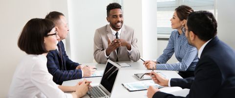 Five business pros conferring in office room with laptop open and notepads on tabletop. 