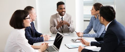 Five business pros conferring in office room with laptop open and notepads on tabletop.  