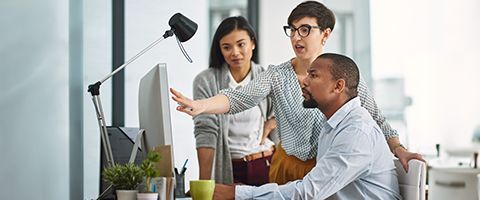 Two colleagues stand at third person’s desk while one points at monitor as the other two examine the screen. 