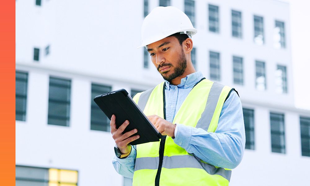 Construction worker wearing a white hard hat and neon yellow safety vest, using a tablet device outdoors in front of a modern building.