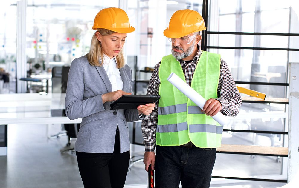 Two construction professionals wearing yellow hard hats stand in a modern office setting. One person is holding a tablet and appears to be discussing plans while the other is holding rolled-up blueprints.