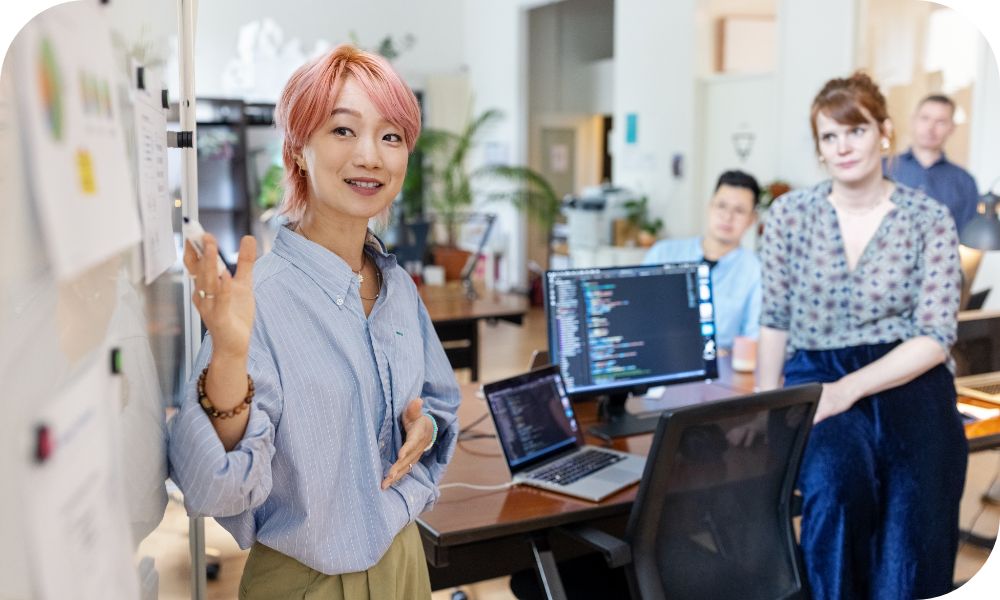 Woman with pink hair presenting in front of three other people sitting at a row of desks and laptops