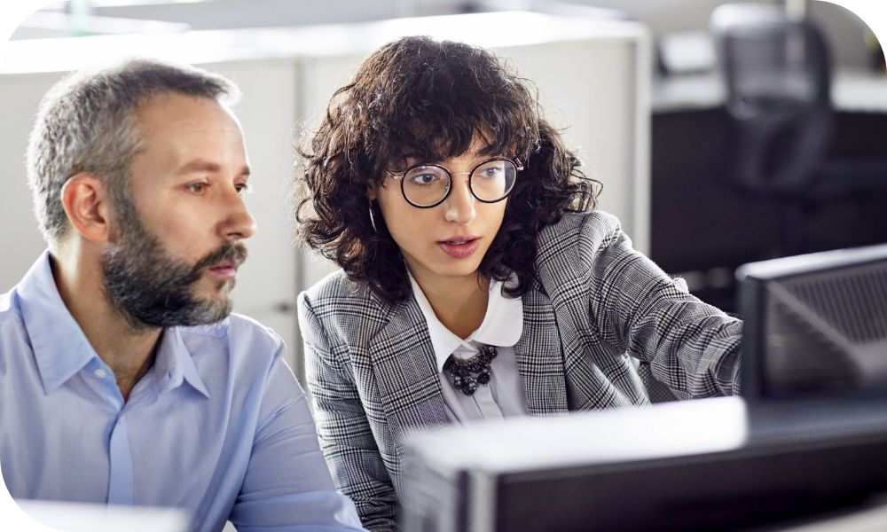 Man and woman sitting in a row of desks while working on a desktop monitor in front of them