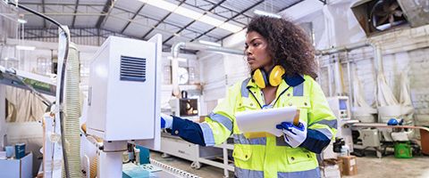 A person in brightly colored protective clothing holds a tablet device in one hand while entering information on a piece of manufacturing equipment with the other 