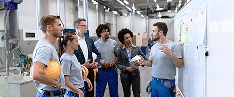 A group of people in a warehouse or factory stand in front of a whiteboard as another person speaks to them