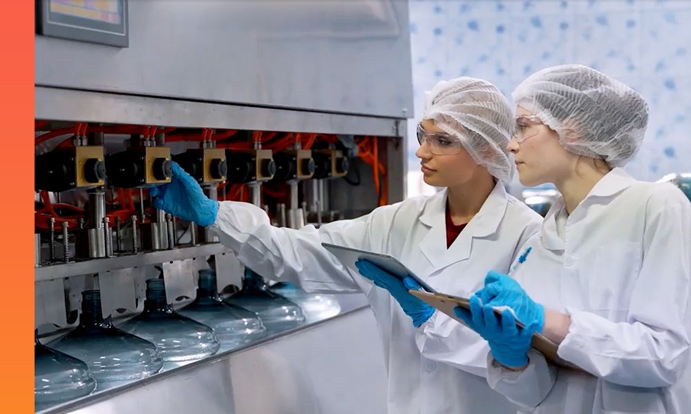 Two people wearing lab coats and safety glasses work on a machine as one holds a tablet and the other takes notes