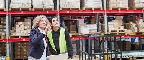 A person in a warehouse with inventory on shelves in the background points up at something as another person looks on 