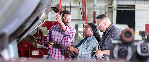 Three people in safety glasses look at equipment in a factory or warehouse