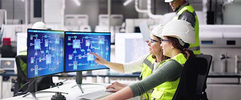 Two people wearing hard hats and safety vests view diagrams on a monitor in a manufacturing facility