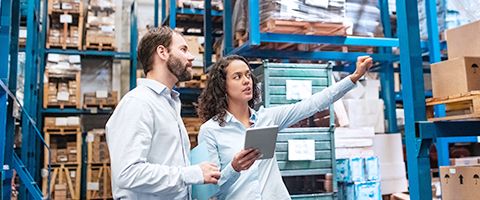 A person in a warehouse holds a tablet in one hand and points up at something with the other hand as another person watches