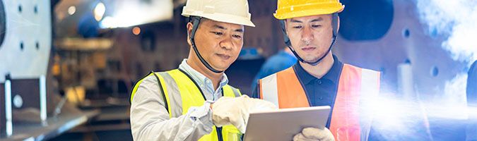 Two workers in yellow hard hats discussing information on a tablet.
