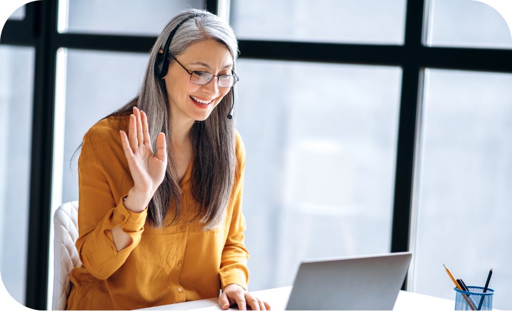 A person sits near a window and talks on a headset while waving into a webcam on a laptop.