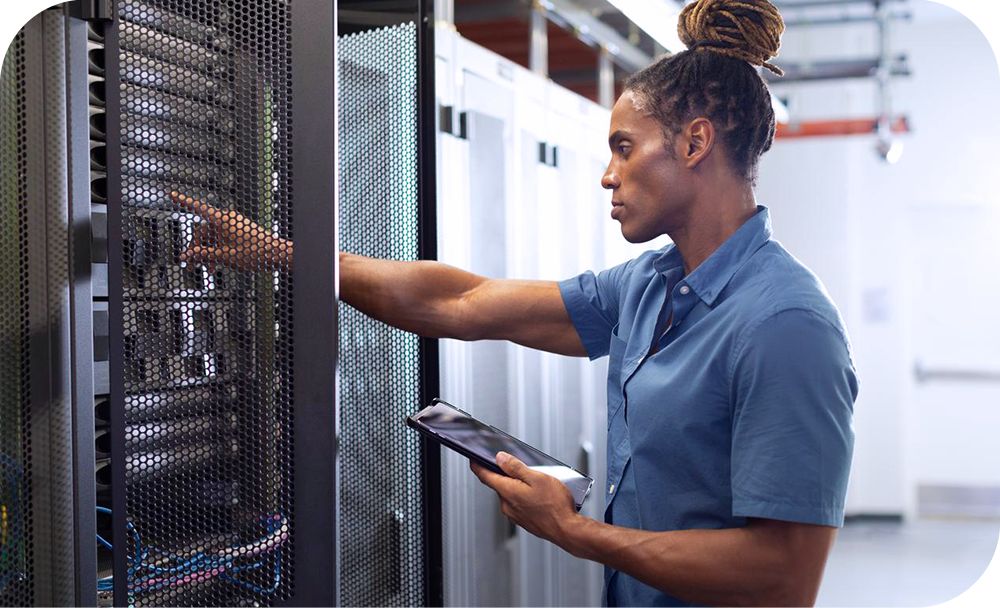 A person in casual business attire holds a tablet device and presses a button on a machine in a server stack