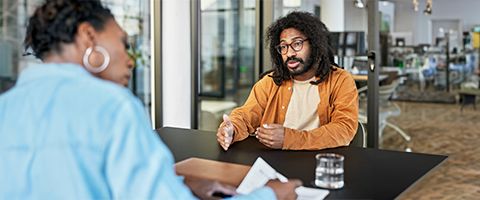 A customer in casual attire talks to a businessperson who is looking at papers on a table