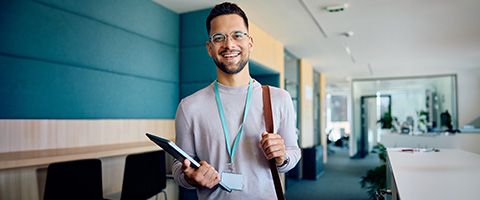 An employee standing in an office holding a laptop in one hand and wearing an ID badge on a lanyard around his neck