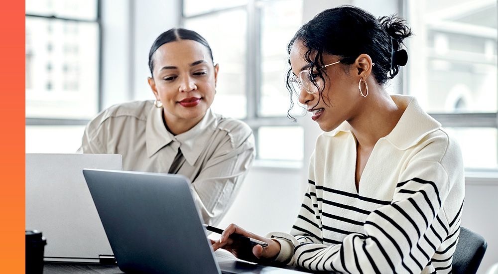 Two professionals collaborating and looking at a laptop screen, sitting side by side in an office environment