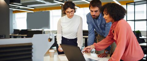 Three people in an office standing and talking over a laptop.