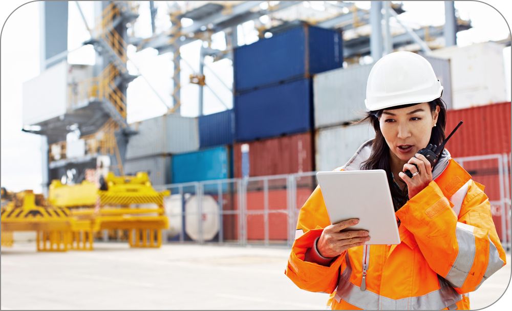 Person in safety gear holding a tablet and walkie talkie in front of storage containers.