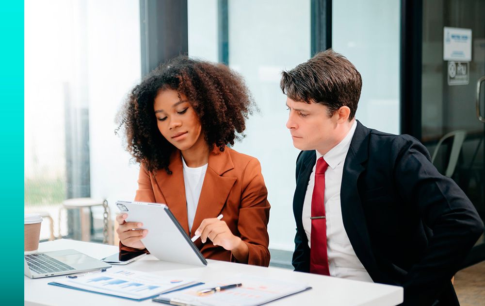 Two people in business dress sitting at a desk in an office looking at information on a tablet