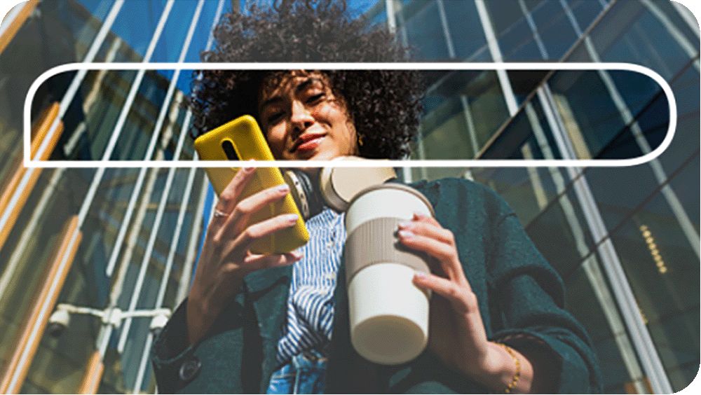 Smiling woman near high-rise buildings looking at her mobile phone and holding a cup of coffee.