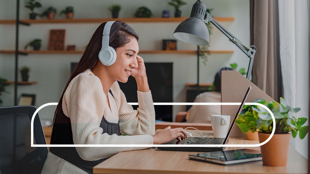 Woman wearing headphones sitting at a desk in a home office with a laptop and tablet in front of her