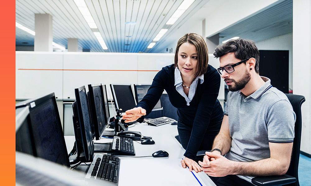 Female coworking standing and pointing at row of monitors on desk while male colleague studies the data on the screen. 