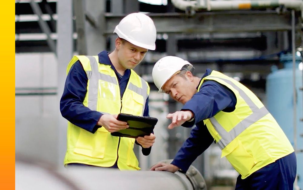 Two men in vests and hard hats work outside with a tablet. 