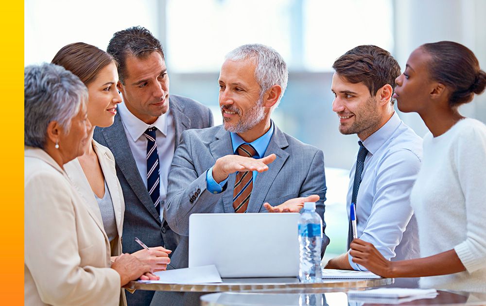 A group of professionals gather around a tall, round table to look at and discuss information on a laptop