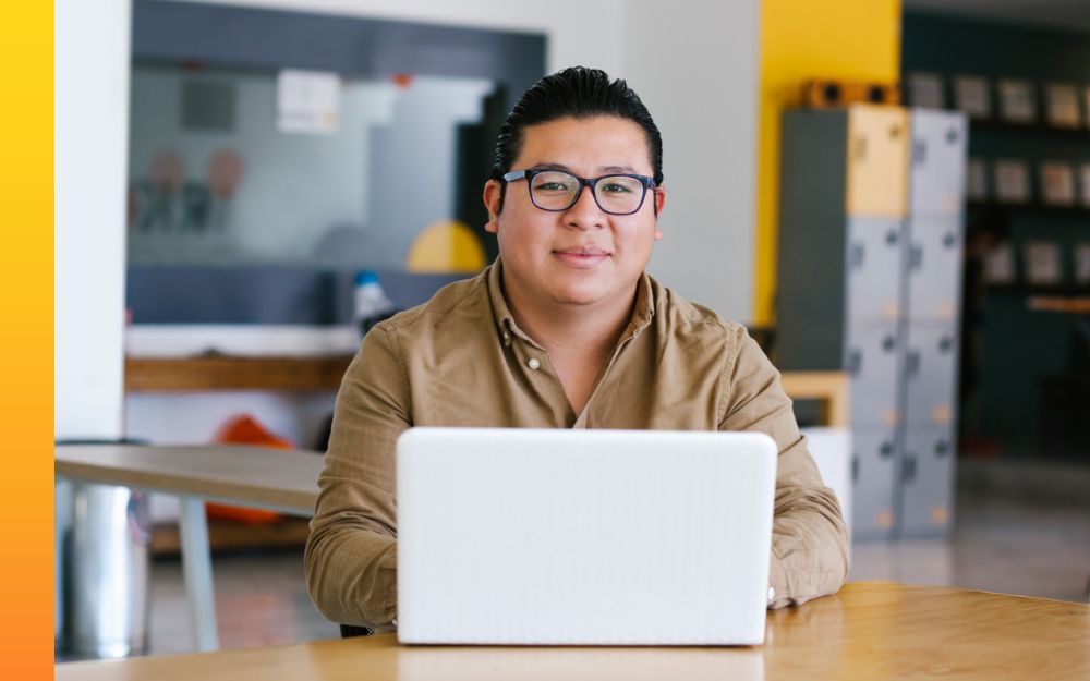 A person in glasses works on a laptop while seated at a table in a school.