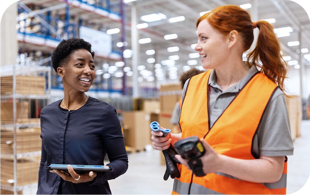 Two people talking in a warehouse setting, one wearing a safety vest and holding a device and the other holding a smart tablet.