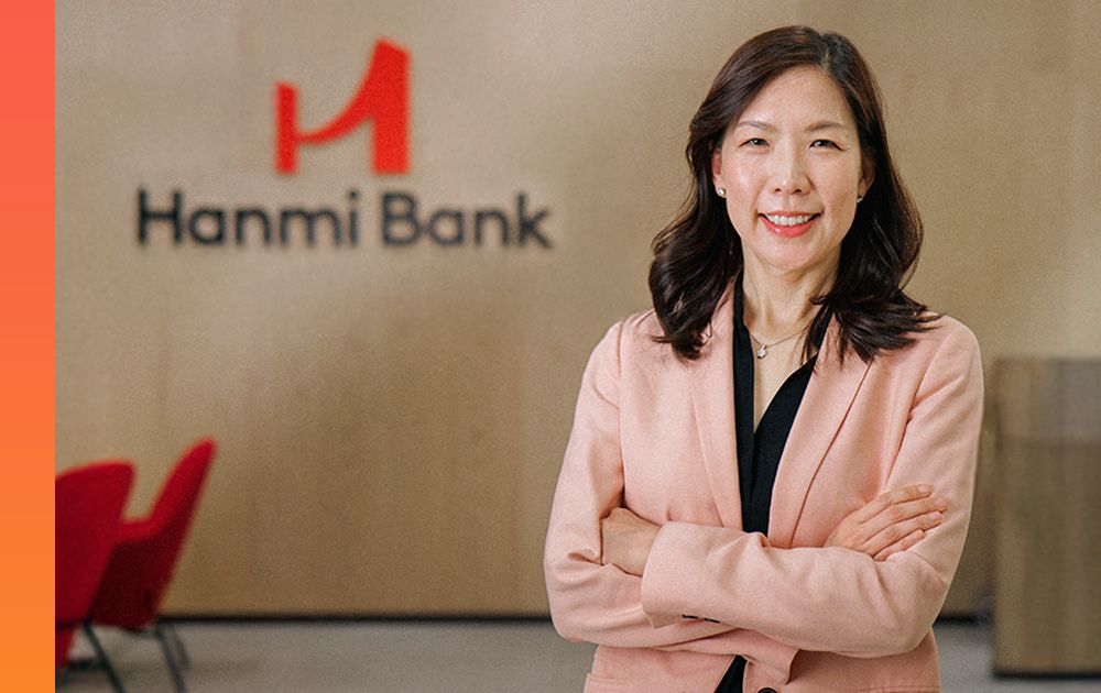 Smiling banker in a light pink blazer, arms crossed, standing in front of a Hanmi Bank logo.