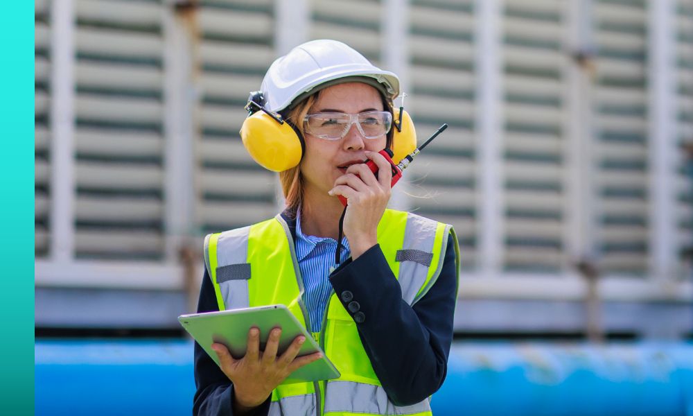 Worker in a yellow safety vest and white hard hat holding a tablet while talking into a two-way radio