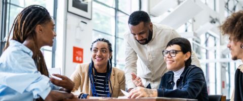 A group of students, smiling and conversing, sit together at a table to work on studies