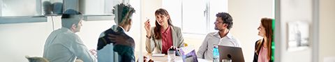 A businessperson confidently leads a conversion during a meeting at a table with four other coworkers