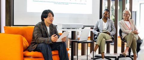 An individual sits in a bright orange chair and presents on a projector using their tablet while professionals sit nearby as a panel and listen