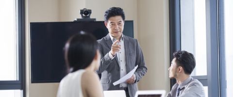 Person in business attire standing in front of a conference table speaking to others seated at the table. 