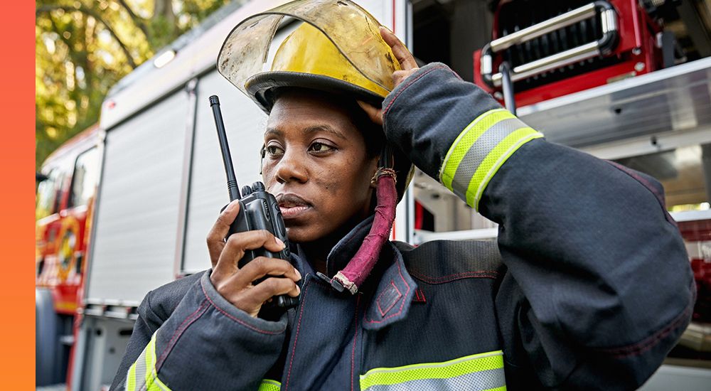 Person in firefighter attire standing next to a firetruck and talking into a handheld device.