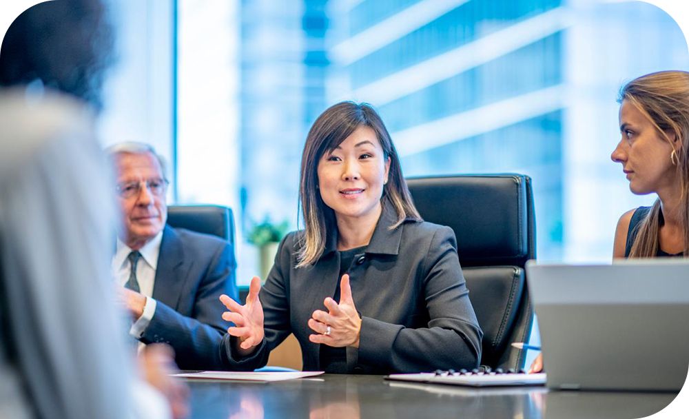 A professional uses their hands to gesture while talking with coworkers in a meeting room with a blurred building in the background