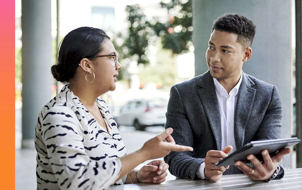 Two businesspeople converse while referencing information on a tablet 