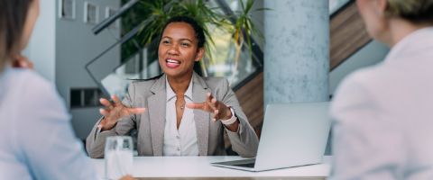 Businessperson sitting at a table speaking and gesturing to two people facing them 