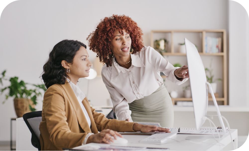 Two people collaborating on a project, with one seated and the other standing, pointing at a computer screen in a room with shelves in the background