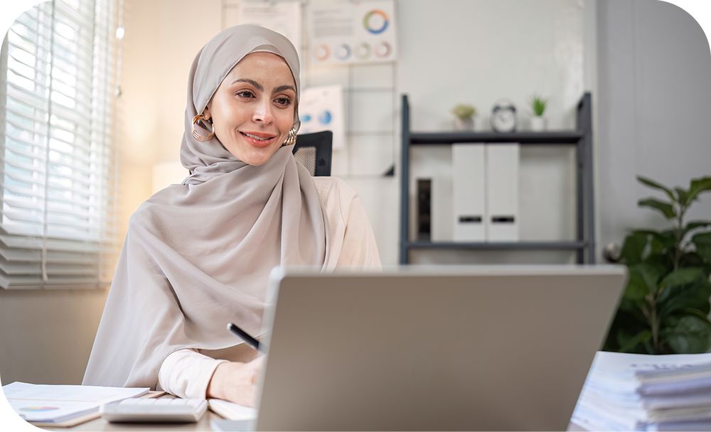 Person in beige sitting at a desk writing on a notepad and smiling at a laptop.