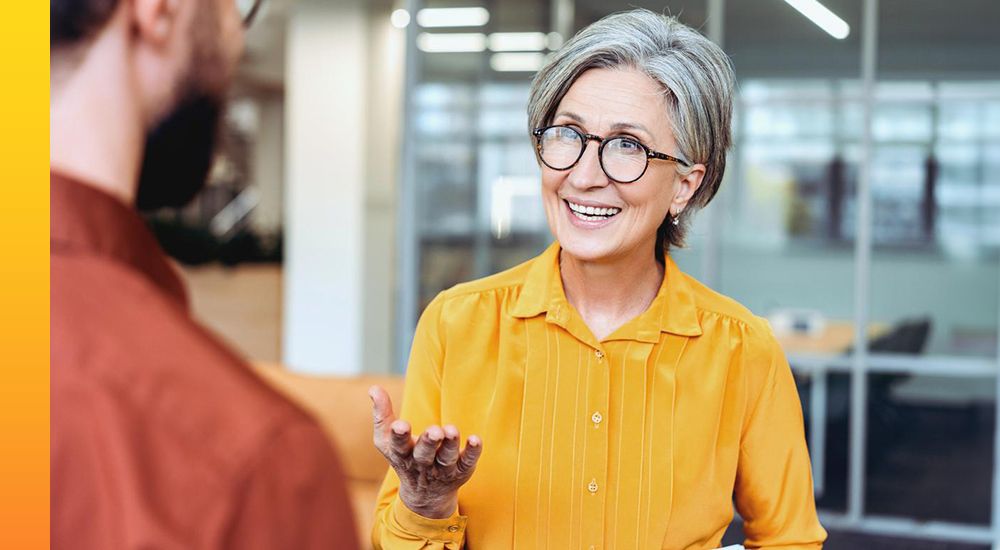 Person in yellow standing in an office, smiling and talking to another person.