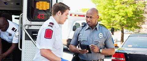 EMS first responder standing behind opened ambulance talking with police officer writing info on computer tablet.