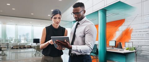 Two colleagues standing together in a modern office, reviewing information on a tablet, with teal and orange walls and open workspaces in the background.