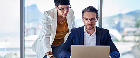 Two colleagues collaborating at a desk in a bright office, one seated and working on a laptop while the other stands nearby, leaning in to review the screen together