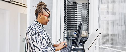 A person standing in a data center, typing on a laptop beside tall server racks, wearing a patterned blouse and working among network and computing equipment.