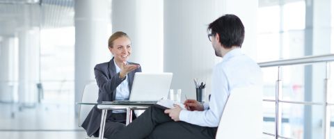 Two people in business attire sitting at a desk and having a discussion over a laptop.